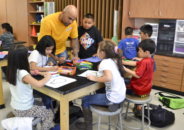 Kinder sitzen um einen Klassentisch mit Papieren, Stiften und Taschen, während ein Mann davorsteht, mit Schränken und Regalen im Hintergrund.