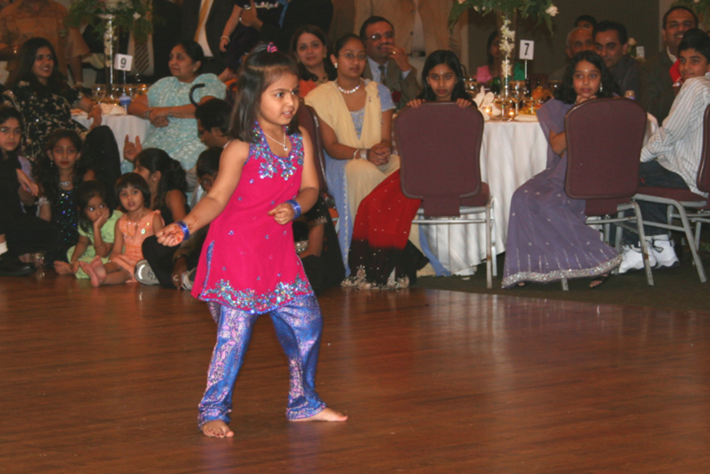 Ein Mädchen tanzt im Vordergrund auf einem Holzboden, während im Hintergrund viele Menschen an Tischen mit weißen Tischdecken und kleinen Pflanzen sitzen.