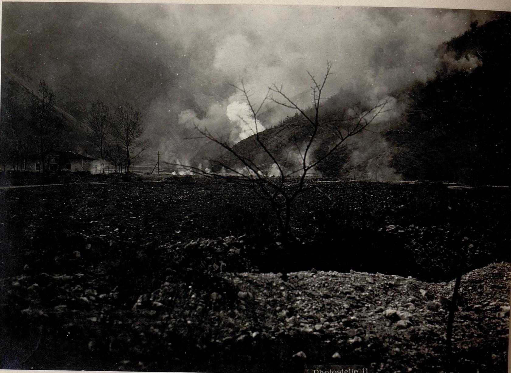 Schwarz-weiß-Foto eines Feuers in einem Feld mit Bäumen, Häusern, Hügeln und Rauch, der in den Himmel steigt; Text unten.