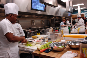Eine Gruppe von Köchen in einer Küche, die Essen auf einem Schneidbrett zubereiten, wobei ein Koch im Vordergrund eine weiße Schürze und eine Mütze trägt und ein Messer hält und von Utensilien, Schüsseln, Gemüse, einem Mobiltelefon, Flaschen und anderen Gegenständen umgeben ist, während im Hintergrund ein Fernseher und Deckenlampen zu sehen sind.
