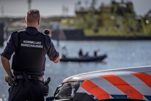 Ein Polizist in Uniform steht neben einem Polizeiwagen in der Nähe eines Gewässers und hält ein Funkgerät in der Hand, mit einem Boot mit ein paar Leuten im Hintergrund und dem Himmel darüber.