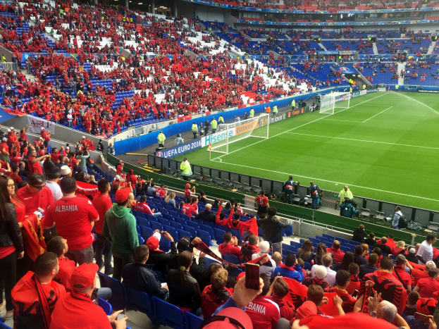 Eine große Menschenmenge in einem Stadion schaut einem Fußballspiel zu, mit Menschen auf Sitzen und Stehplätzen, Bannern, einem Torpfosten und Deckenleuchten.