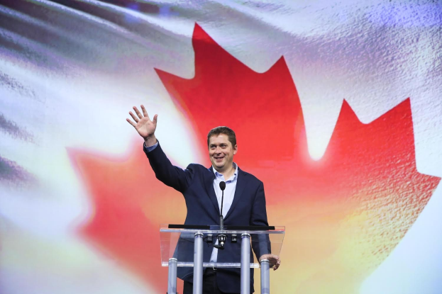 Kanadischer Premierminister Alex Salmond steht an einem Rednerpult mit Mikrofonen, lächelt vor einer kanadischen Flagge und einem Bildschirm.