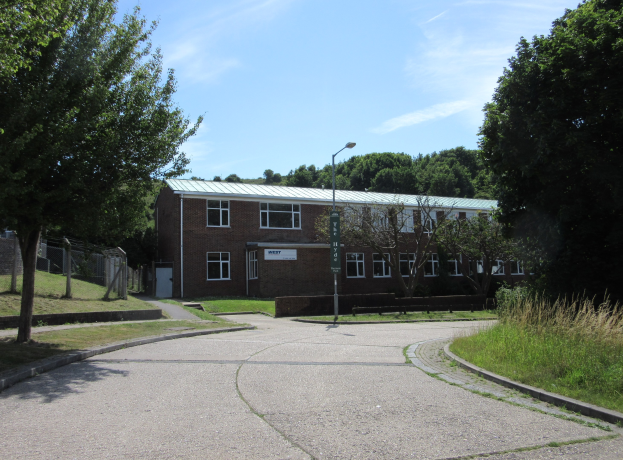 Ein zweistöckiges Schulgebäude mit mehreren Fenstern, einem Straßenmast, einem Schild und einer Baumgruppe, umgeben von saftigem Gras und einem Zaun, unter einem bewölkten Himmel.