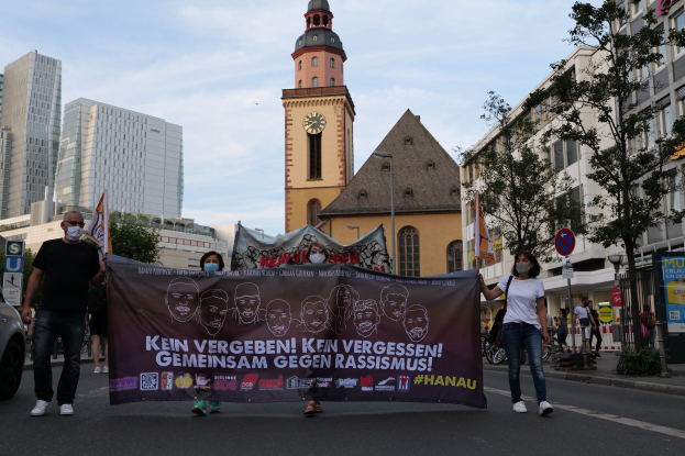Eine Gruppe maskierter Menschen marschiert auf einer Straße und hält ein Banner, daneben steht ein geparktes Auto, im Hintergrund sind Gebäude und Bäume zu sehen sowie ein Uhrenturm unter einem klaren blauen Himmel.