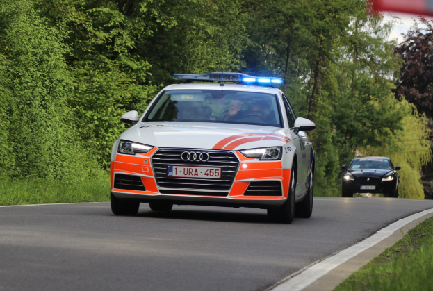 Polizeiauto Audi A4 auf einer grasbewachsenen Straße mit zwei Insassen, umgeben von Bäumen unter einem klaren blauen Himmel.