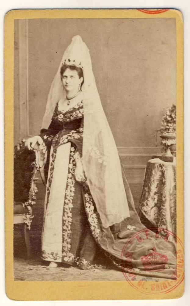 Altes Schwarz-Weiß-Foto einer Frau in traditionellem russischen Hochzeitskleid, das auf einem Stuhl sitzend einen Blumenstrauß hält, mit einem Blumenstrauß auf einem Tisch rechts und Text und einem Stempel unten.