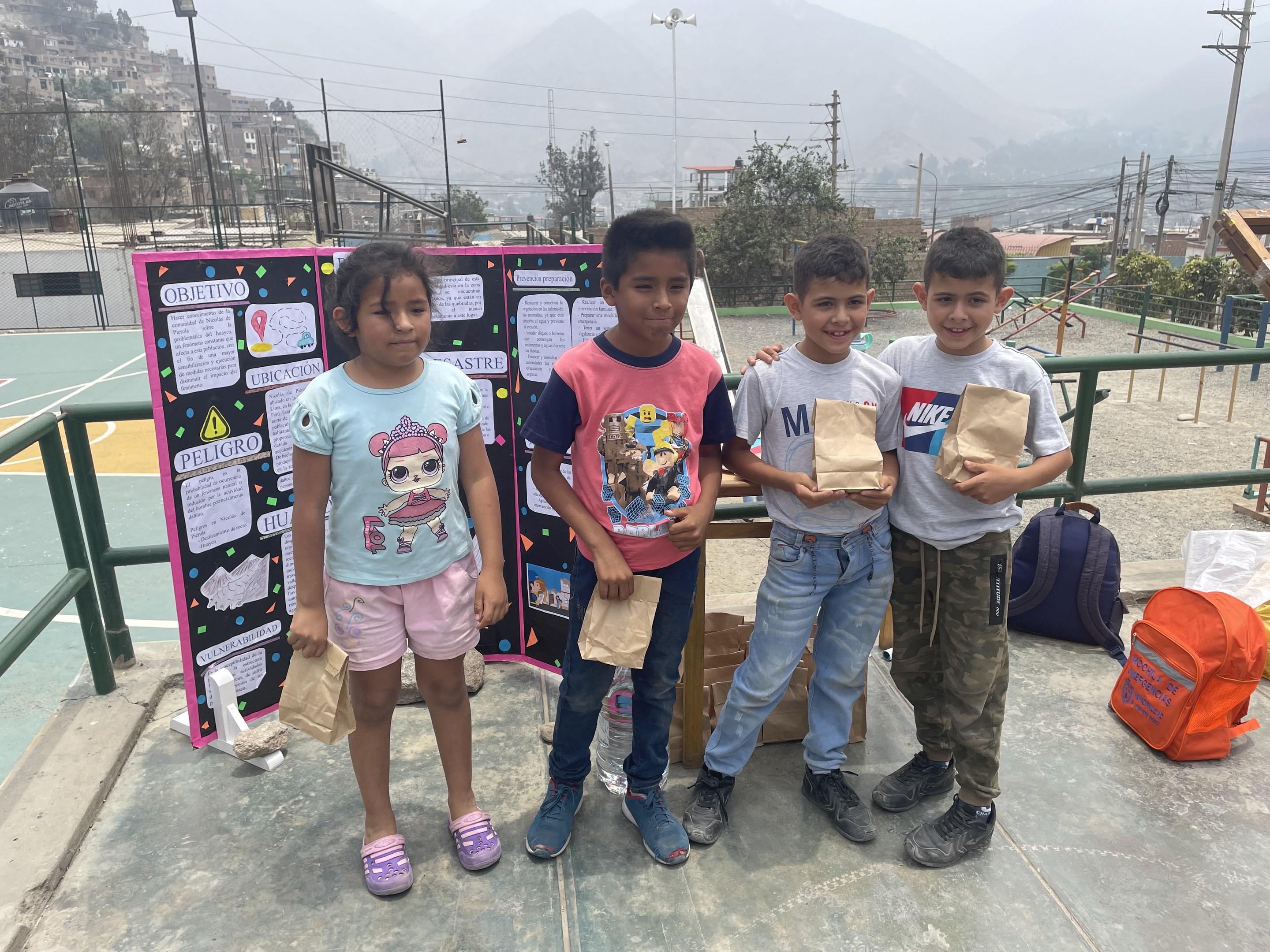 Gruppe von Kindern auf einem Basketballfeld mit Papierbeuteln in den Händen, Gegenstände auf der rechten Seite und eine Tafel mit Text auf der linken Seite, vor einem Hintergrund aus Zäunen, Pfählen, Straßenlaternen, Bäumen, Gebäuden, Hügeln und Himmel.