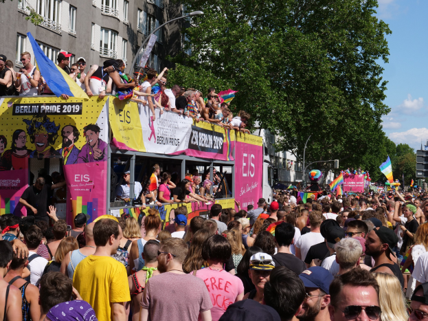 Eine große Menge marschiert während des Pride 2019 in Berlin die Straße entlang, viele tragen Kappen und Schutzbrillen, einige halten Fahnen, mit Bannern, Laternenmasten, Bäumen und Gebäuden im Hintergrund unter einem bewölkten Himmel.