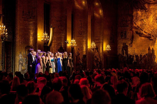 Eine Gruppe von Menschen auf einer hell erleuchteten Bühne im Royal Opera House London mit bemalten Kulissen, während das Publikum von unten zusieht.