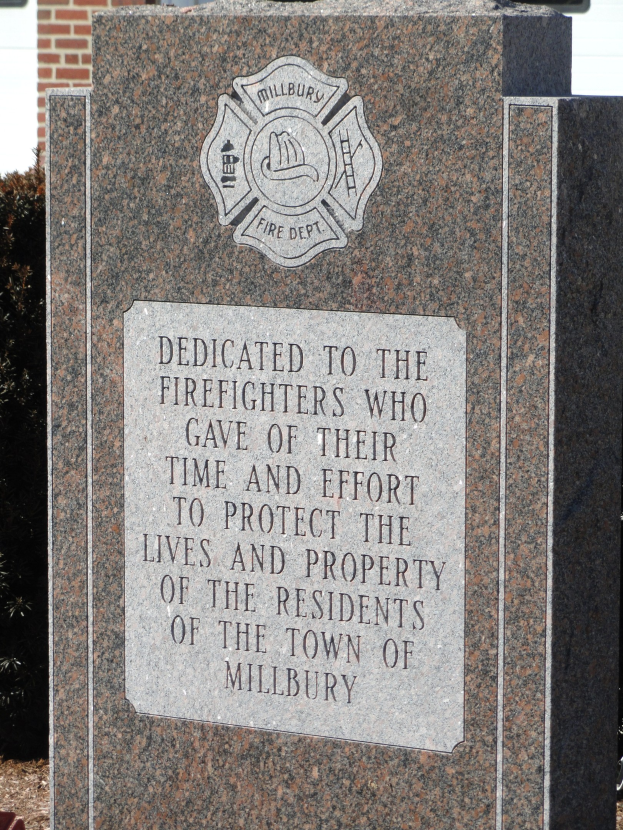 Denkmalstein mit einer Plakette für Feuerwehrleute in Millbury, vor einem Backsteingebäude mit umgebender Vegetation.