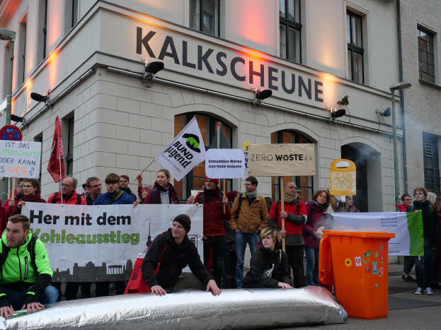 Eine Gruppe von Menschen mit Protestschildern und Plakaten vor einem Gebäude, mit zwei Personen im Vordergrund und einem Müllcontainer auf der rechten Seite.