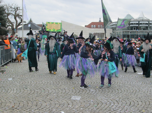 Gruppe von Menschen in Hexenkostümen, die eine Kopfsteinpflasterstraße entlanggehen und schwarze und lila Kleider und Hüte tragen, einige halten Fahnen, mit einem Geländer auf der linken Seite und Gebäuden, Bäumen und einem klaren blauen Himmel im Hintergrund.