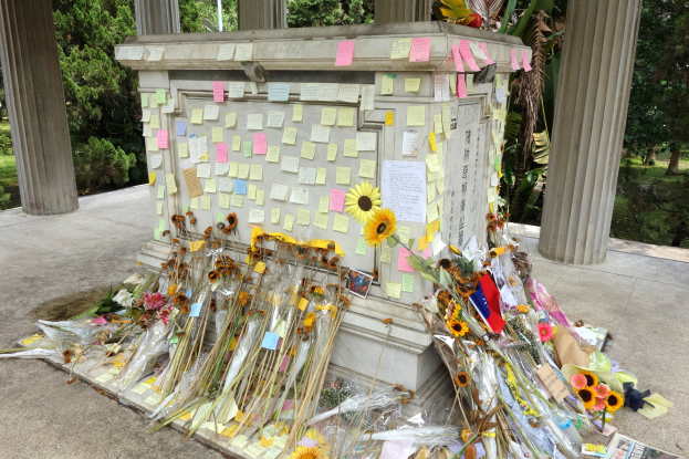 Gedenkfeier mit einem zentralen Pfeiler, der mit Post-it-Notizen und Blumen geschmückt ist, umgeben von Bäumen, Pflanzen und Gras in einer friedlichen Umgebung.