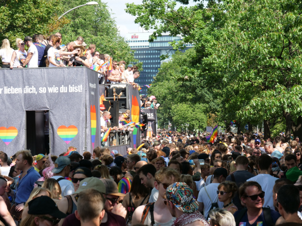 Eine große Menschenmenge steht vor einem Lastwagen bei der Christopher Street Day Parade in Berlin, viele tragen Mützen und Schutzbrillen, einige halten Fahnen, mit einem Banner auf dem Lastwagen, Bäumen und Gebäuden im Hintergrund und einem bewölkten Himmel.