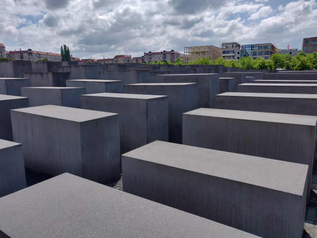 Konkrete rechteckige Blöcke, die in einem Gittermuster am Holocaust-Mahnmal für die ermordeten Juden Europas in Berlin angeordnet sind, mit Bäumen, Gebäuden und Wolken im Hintergrund.