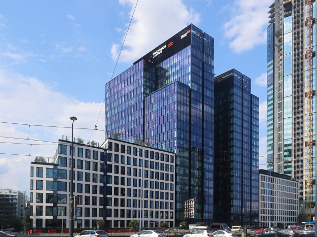 Ein großes mehrstöckiges Gebäude mit zahlreichen Fenstern, identifiziert als die Deutsche Bank-Zentrale in Frankfurt, Deutschland, in einer städtischen Umgebung mit Straßeninfrastruktur, Fahrzeugen, Grünflächen und einem bewölkten Himmel.