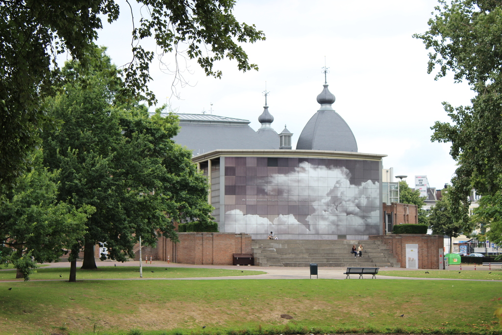 Ein Palast mit Gebäuden dahinter, umgeben von Grasland mit Bäumen auf beiden Seiten und im Hintergrund, unter einem klaren Himmel.