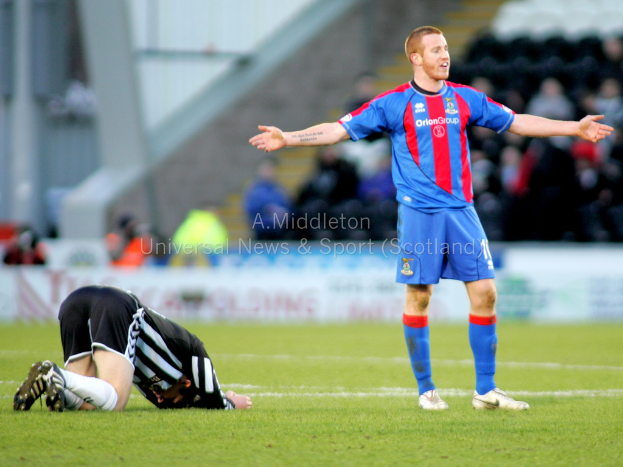 Ein Stadion mit einer Person, die steht, und einer anderen, die auf dem Rasen kniet, sowie einer Gruppe von Menschen im Hintergrund, die Schilder mit Text halten.