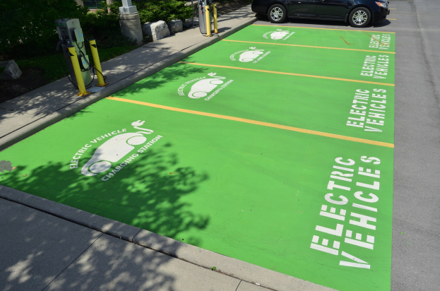 Elektroauto auf einem Ladeplatz mit einer grünen Bodenmatte mit Text und Bildern in einer Parkanlage umgeben von Bäumen, Pflanzen, Pfählen und anderen Gegenständen.