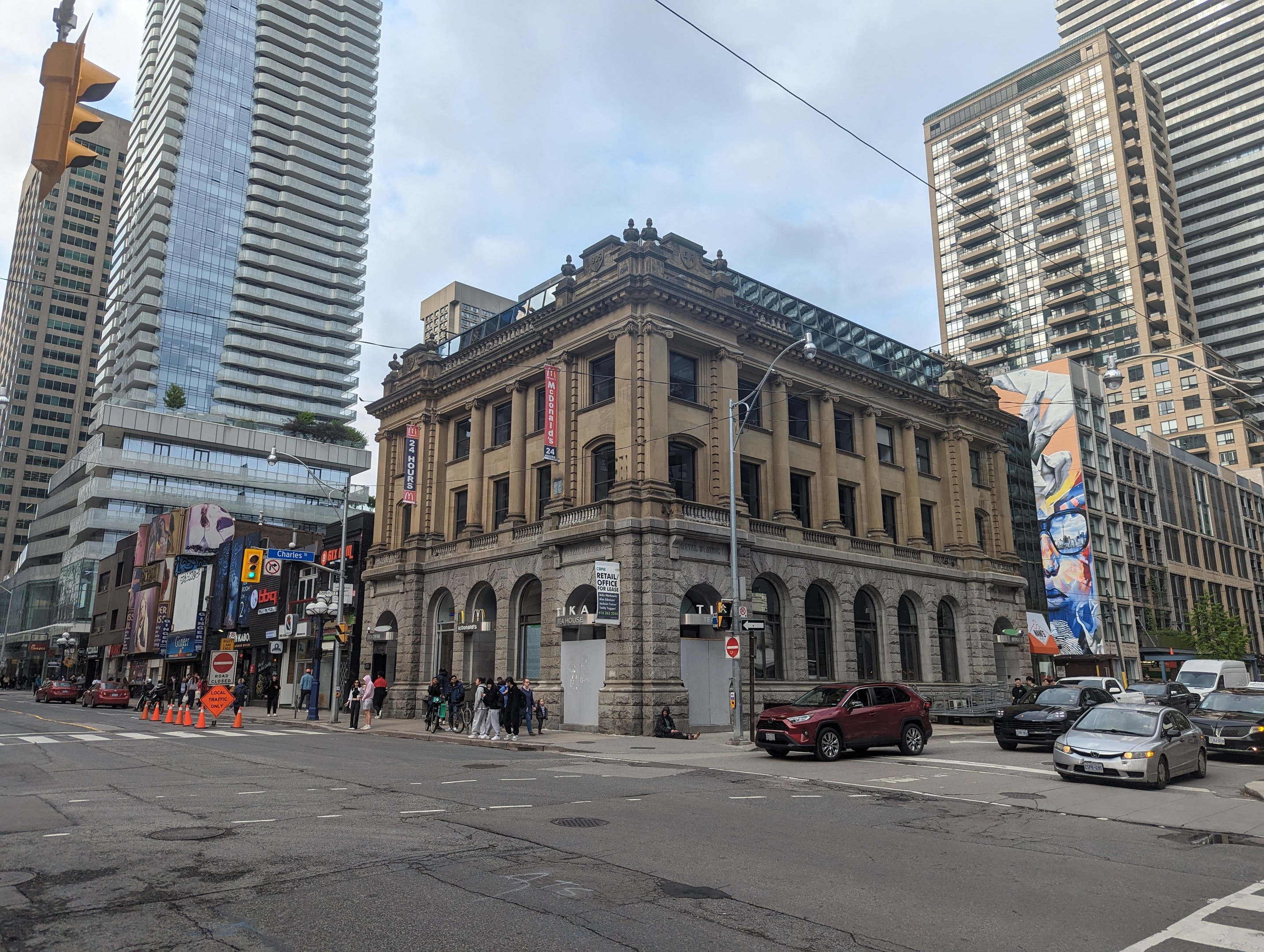 Eine belebte Stadtstraße mit hohen Gebäuden, Fahrzeugen, Fußgängern, Verkehrsinfrastruktur, Bäumen und der Toronto Public Library als Mittelpunkt unter einem bewölkten Himmel.