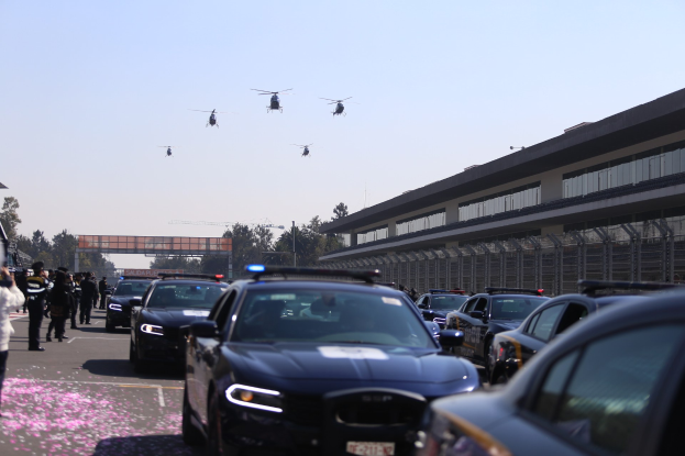 Polizeifahrzeuge vor einem Gebäude mit Menschen auf der linken Seite, Bäumen, einer Brücke und vier Hubschraubern am Himmel.