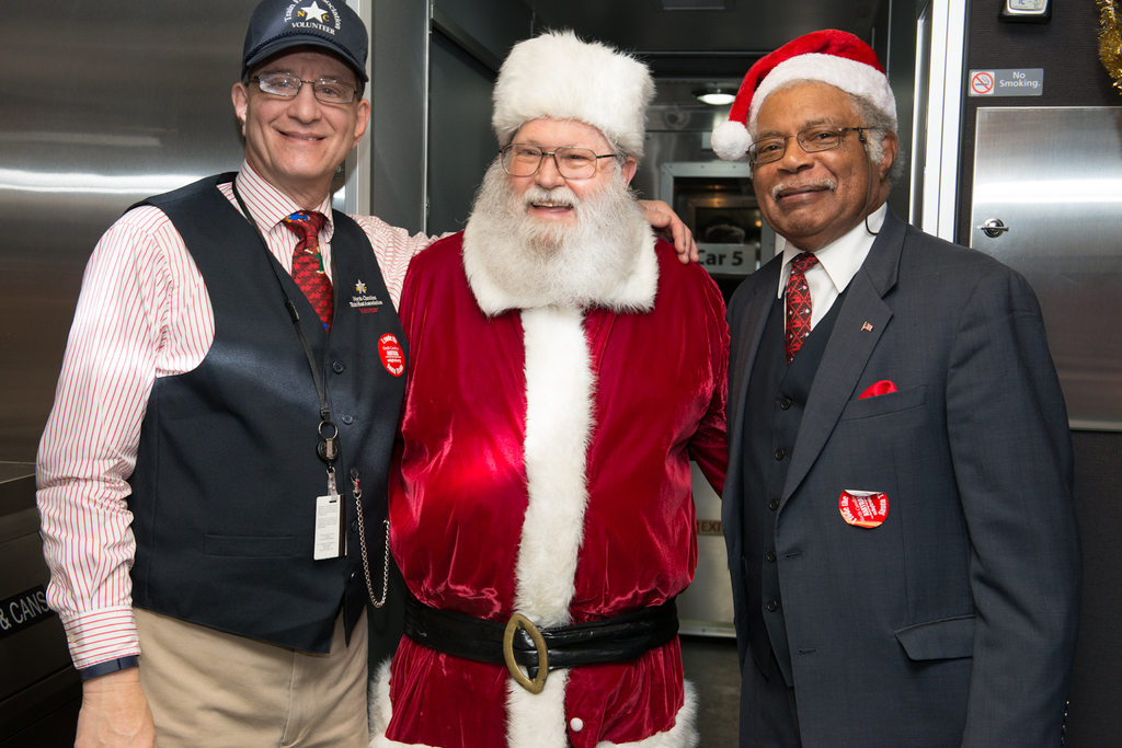 Eine Gruppe von Menschen mit Brillen, Mützen, Krawatten, Ausweisen und Abzeichen, darunter eine Person in einem Weihnachtskostüm, vor einer Stahlwand mit Lichtern, Schildern und anderen Gegenständen im Hintergrund.