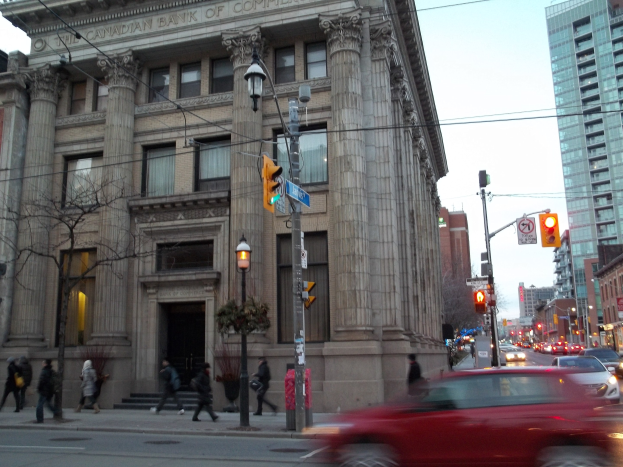 Eine belebte Stadtstraße mit Fahrzeugen, Fußgängern, Verkehrszeichen, Schildern, Bäumen und Gebäuden unter einem sichtbaren Himmel, mit einem roten Auto im Vordergrund.