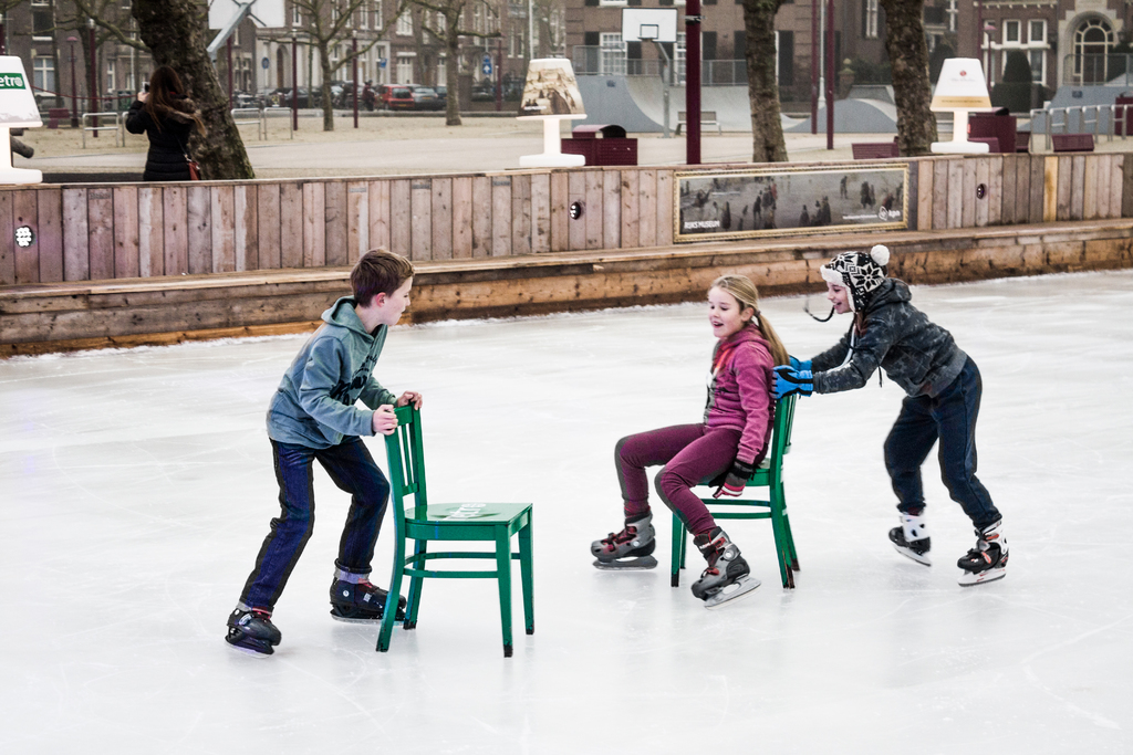 Kinder beim Skifahren vor einem Spielplatz mit drei Kindern und zwei Stühlen in der Mitte sowie Gebäuden, Bäumen, Bänken, Pfosten und einem Basketballfeld im Hintergrund.