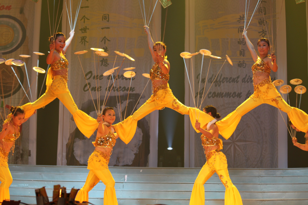 Eine Gruppe von Frauen in gelben Outfits führt Akrobatik auf einer Bühne durch, einige halten Gegenstände, mit einem Publikum und einer textbedeckten Wand im Hintergrund.