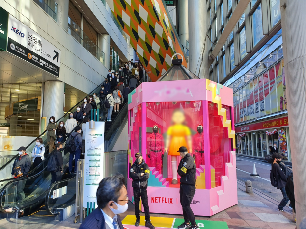Eine Gruppe von Menschen steht vor einem Gebäude mit Glaswänden, Säulen und Texttafeln, einige tragen Masken, während eine Rolltreppe mit Menschen zu sehen ist. In der Mitte steht ein pinker Kasten mit Schaufensterpuppen und der Aufschrift "Netflix's new Netflix booth in Tokyo".