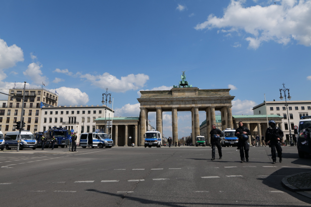 Gruppe von Polizisten vor dem Brandenburger Tor in Berlin, Deutschland, mit den Säulen und der Statue des Tors, umgeben von Gebäuden, Fahrzeugen, Laternenmasten, Verkehrsampeln und einem bewölkten Himmel.