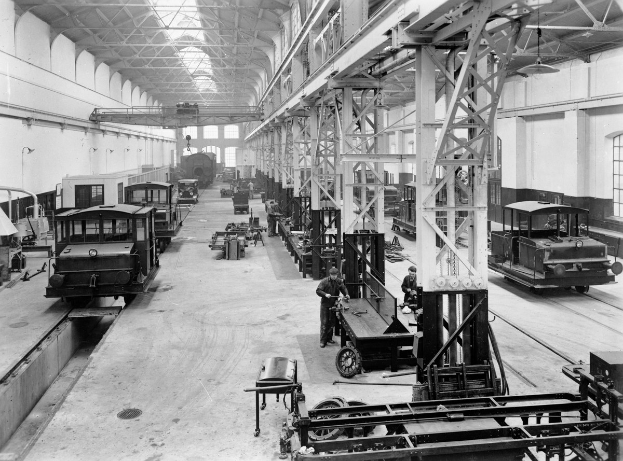 Schwarzes und weißes Foto einer Fabrikhalle mit mehreren reparierten Zügen, Arbeitern, Metallstangen, Deckenlampen und Wandfenstern.