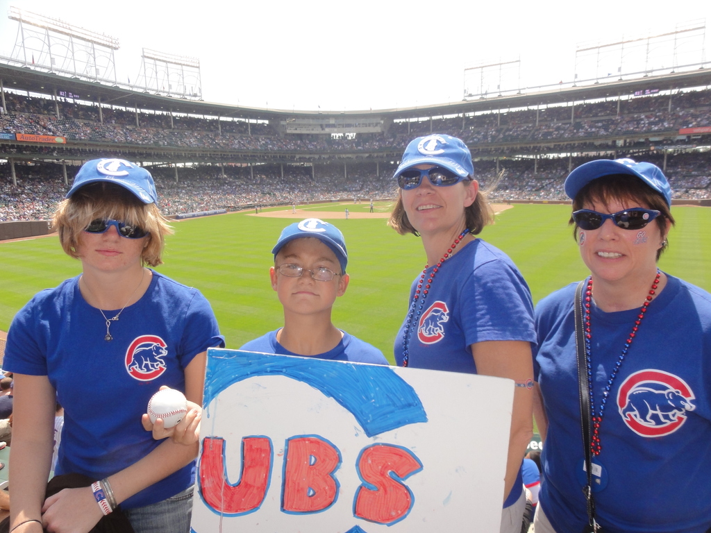Drei Frauen mit Kappen und Brillen, ein Junge mit Brille und Kappe, der einen Ball und ein Schild hält, in einem Stadion mit Publikum und Feld dahinter, unter einem Himmel mit ein paar Lichtern.