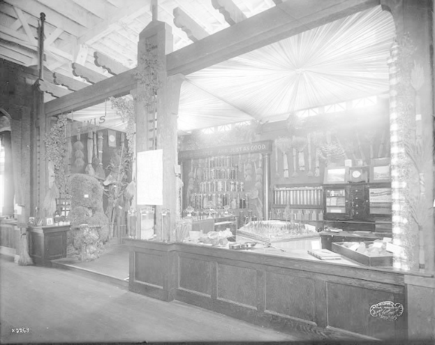 Schwarz-weiß-Foto eines alten Apothekenladens mit einem zentralen Tresen, auf dem verschiedene Gegenstände ausgebreitet sind, Regalen im Hintergrund mit Flaschen und Artikeln gefüllt und Text unten, der darauf hinweist, dass es sich um eine Apotheke handelt.