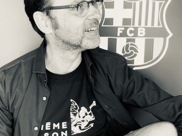 Ein Mann in Brille und schwarzem Hemd lächelt, während er vor einem schwarz-weißen FC Barcelona-Logo sitzt.