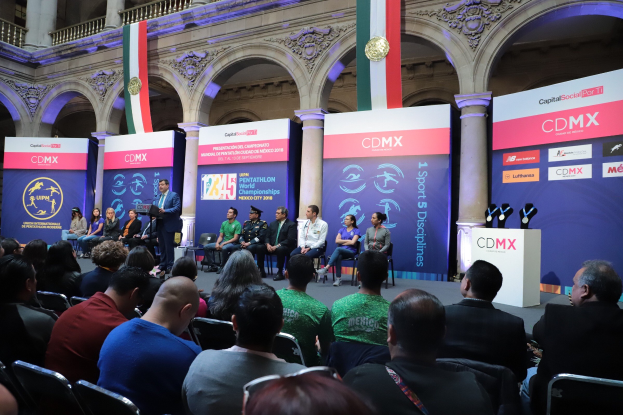 Eine Gruppe von Menschen sitzt in Stühlen vor einer Bühne, auf der ein Mann neben einem Mikrofonständer steht, mit Schildern "Pentathlon-Weltmeisterschaften 2018" hinter der Bühne.