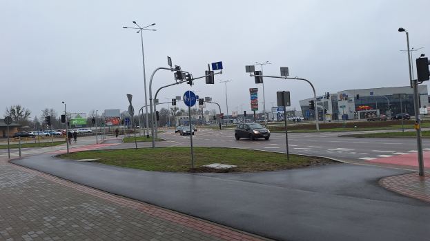 Eine Stadtstraße mit Autos, Gras, Laternen, Verkehrszeichen, Schildern, Bäumen, Gebäuden und einem klaren blauen Himmel.