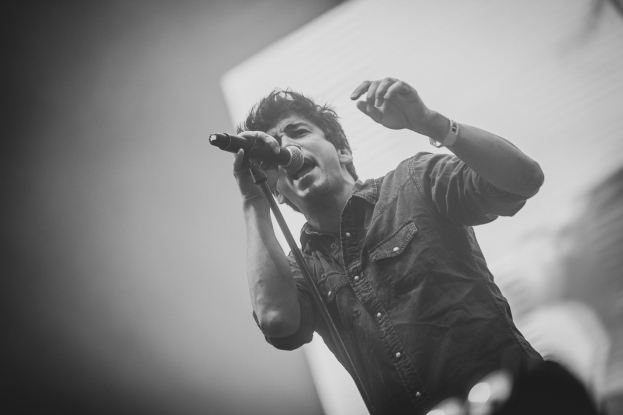 Schwarzes und weißes Foto eines Mannes, der in ein Mikrofon auf der Bühne singt, mit einem unscharfen Gesicht im Hintergrund.