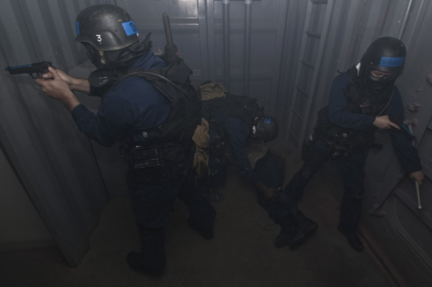 Eine Gruppe von Polizisten in Einsatzausrüstung steht zusammen, einer hölt eine Waffe, mit einem Metallbehälter und einer Leiter im Hintergrund.