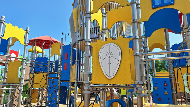 Ein Spielplatz mit buntem Spielgerät, einschließlich Stangen, Polen und einem Schirm, einige Menschen, Bäume und einen klaren blauen Himmel.