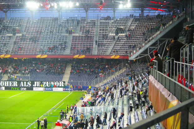Großes Stadion voller Zuschauer bei einem Fußballspiel, mit sitzenden und stehenden Personen, einem Geländer, einer rechten Banner und einem leeren Abschnitt im Hintergrund mit Deckenbeleuchtung.