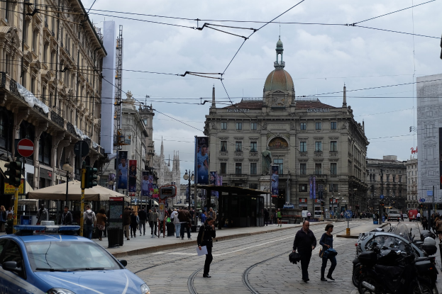 Eine belebte Straße mit einem geparkten Polizeiwagen, Fördernde mit Taschen, fahrende Fahrzeuge, Gebäude mit Fenstern und Plakaten, Laternenmäste, Verkehrsampeln und ein bewölkter Himmel.