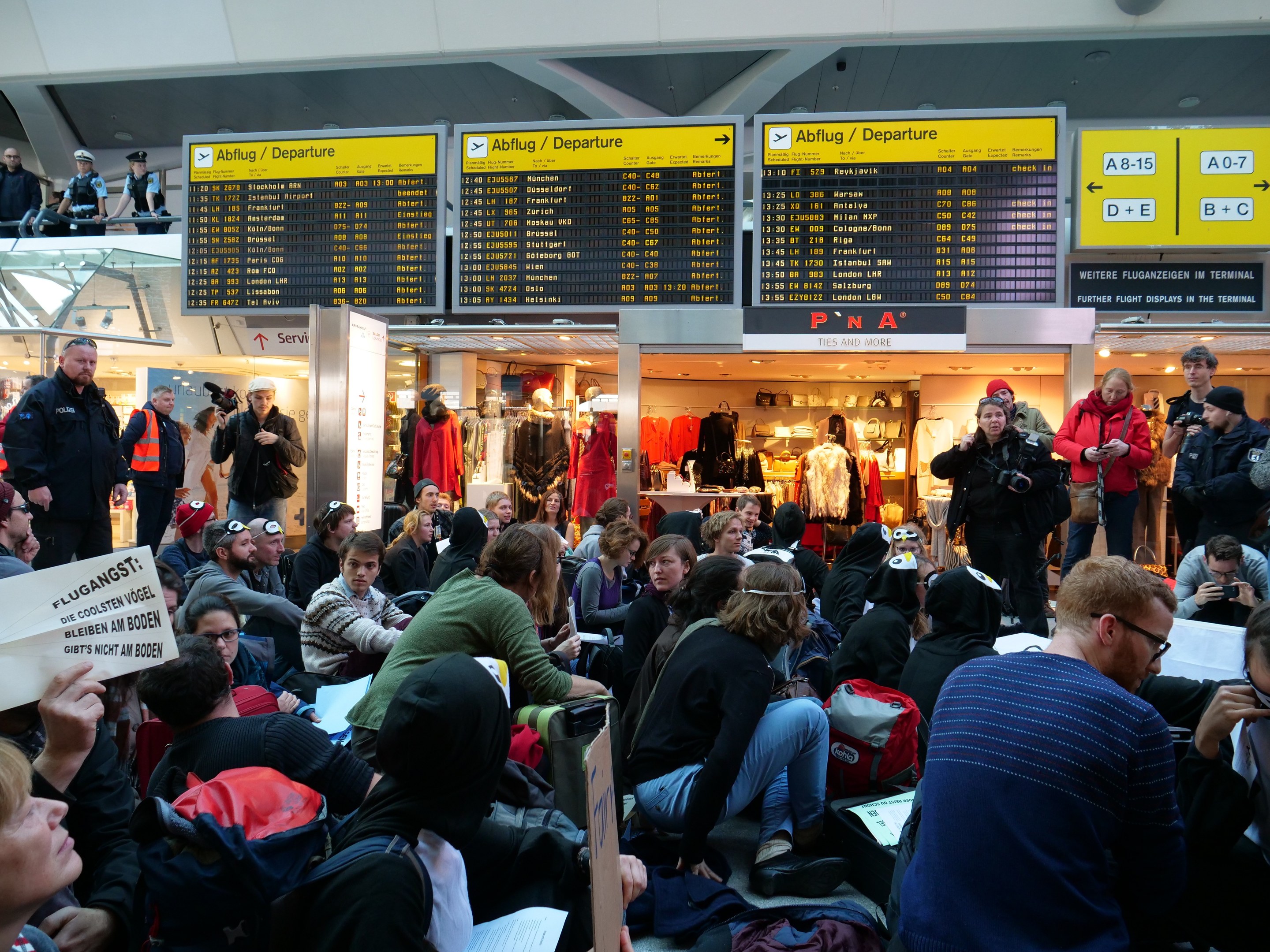 Menschen sitzen und stehen in einem Flughafen während einer Demonstration, mit Informationsschildern, Schaufensterpuppen und Deckenbeleuchtung im Hintergrund.