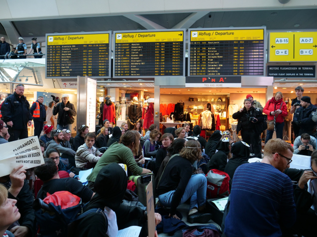 Menschen sitzen und stehen in einem Flughafen während einer Demonstration, mit Informationsschildern, Schaufensterpuppen und Deckenbeleuchtung im Hintergrund.