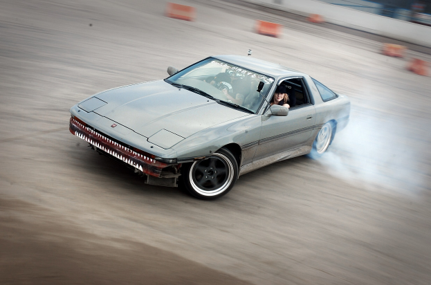 Eine Person in einem Helm fährt ein Auto auf einer Strecke, aus dem Rauch aufsteigt, umgeben von orangefarbenen Gegenständen, mit einem unscharfen Hintergrund, der hohe Geschwindigkeit andeutet.