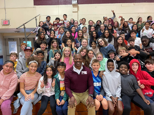 Schüler posieren für ein Foto in einer Turnhalle, einige sitzen auf der Treppe und andere stehen, eine Tür und ein Ausgangsschild sind links zu sehen.
