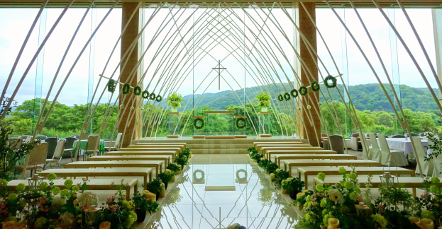 Eine Hochzeitszeremonie in einem großen Saal mit Tischen in Halbkreisform, dekoriert mit Blumen, unter einem klaren Himmel mit Bäumen und Hügeln im Hintergrund.