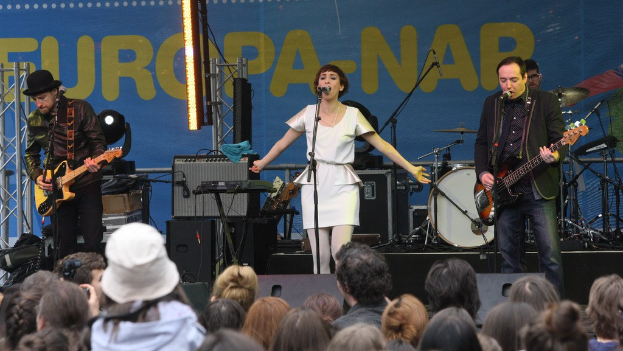 Vier Personen auf der Bühne: eine Frau in einem weißen Kleid, die in der Mitte singt, flankiert von zwei Männern, die Gitarren spielen, mit einer Menge unten und Lautsprechern und einer Wand im Hintergrund.