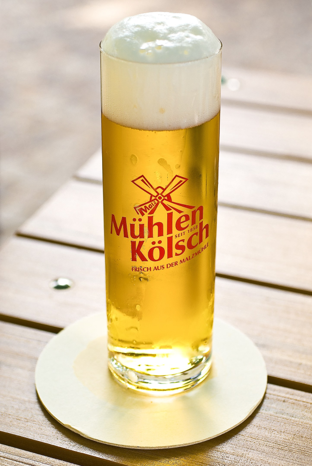 Ein Glas goldfarbenes Münchner Kölsch-Bier, randvoll mit sichtbarer Schaumkrone, steht auf einem hölzernen Tisch mit leicht unscharfem Hintergrund.